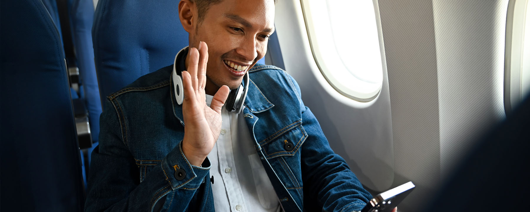 Man in airplane window seat with headphones around neck waving at his phone screen