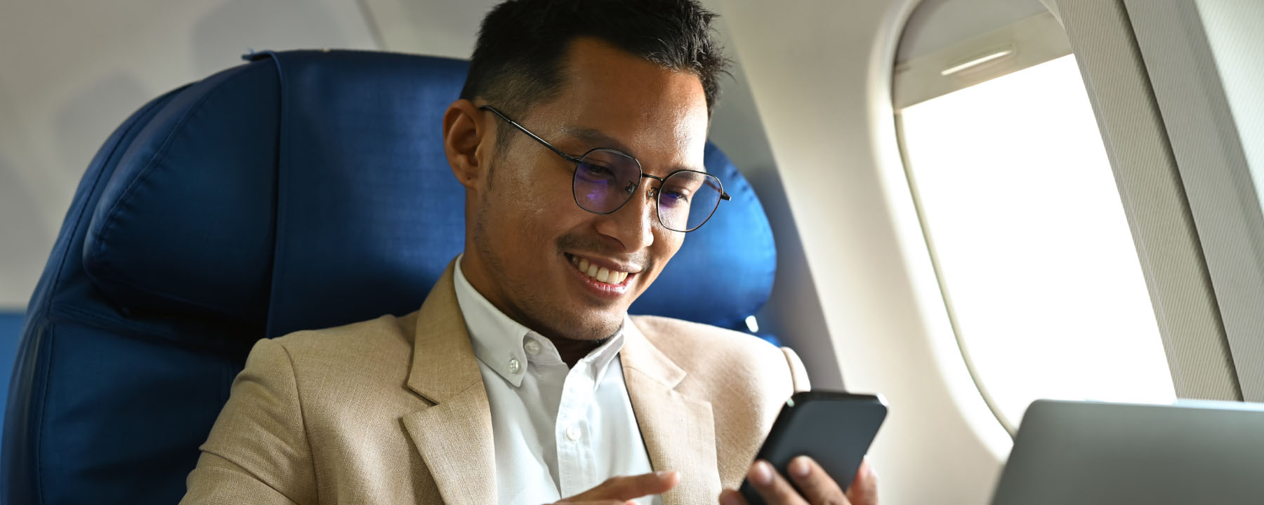 Person in a blazer using a smartphone, seated in an airplane with a laptop open in front of them