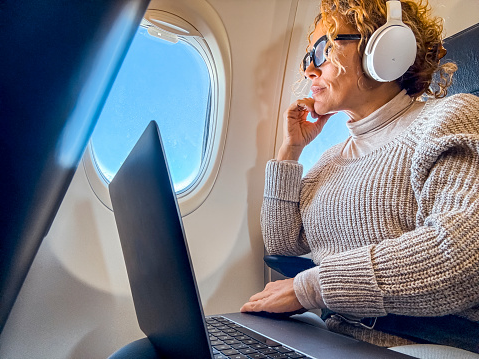 Person wearing headphones and sunglasses, looking out an airplane window, with a laptop on their lap