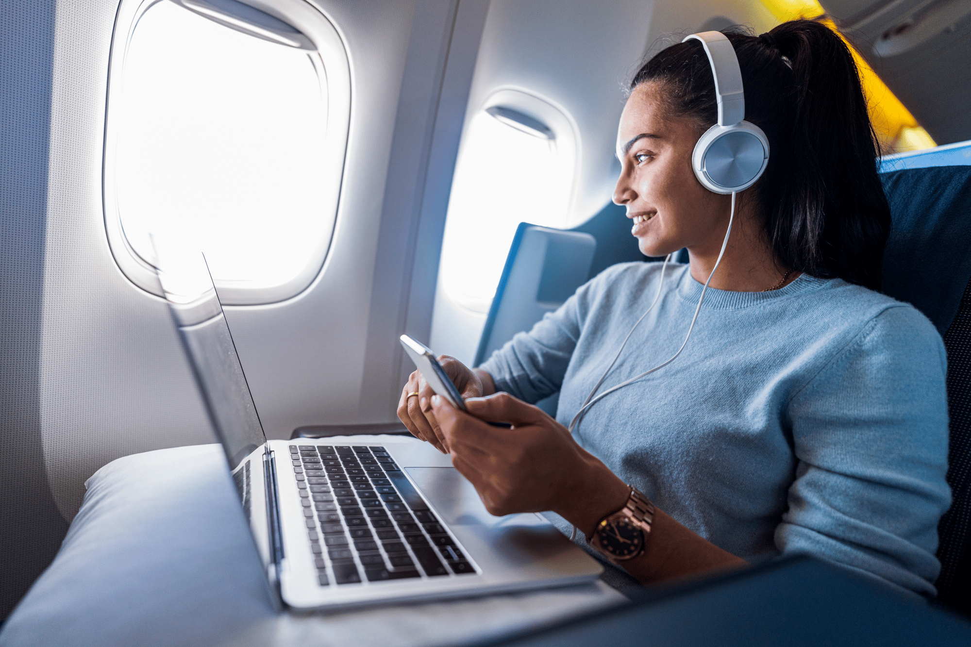 Woman wearing headphones, holding a smartphone, and looking at a laptop on an airplane