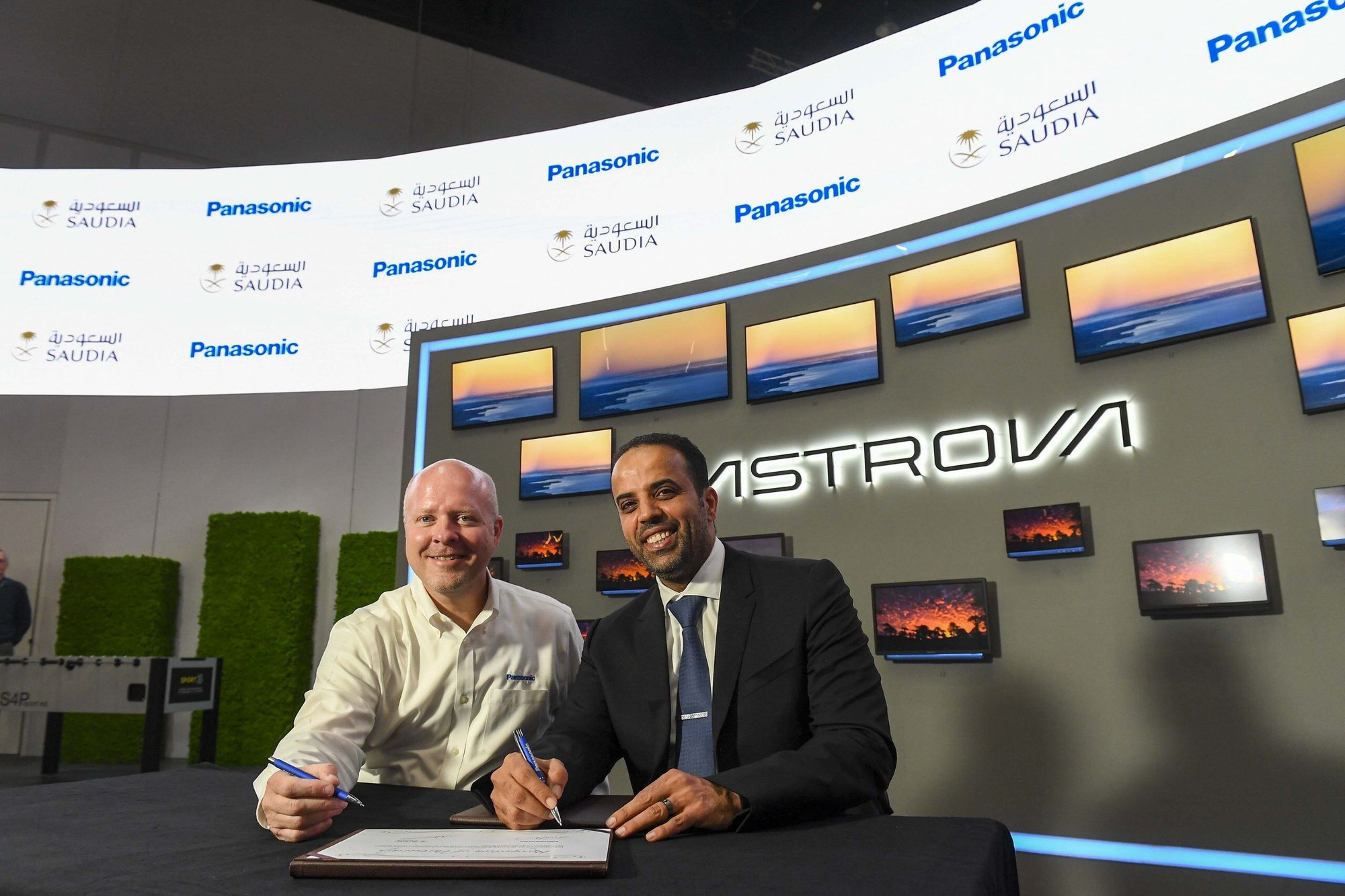 Two men smiling, holding pens, about to sign a document; sunset screens with "Panasonic" and "Saudia" logos