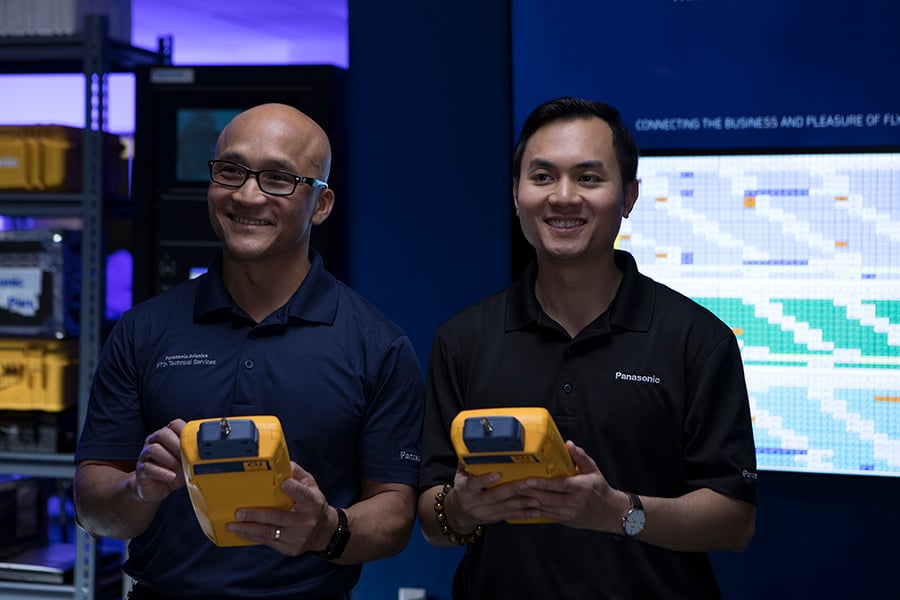 Two men in dark shirts with "Panasonic" logos hold yellow devices, standing before shelves and a screen showing a grid